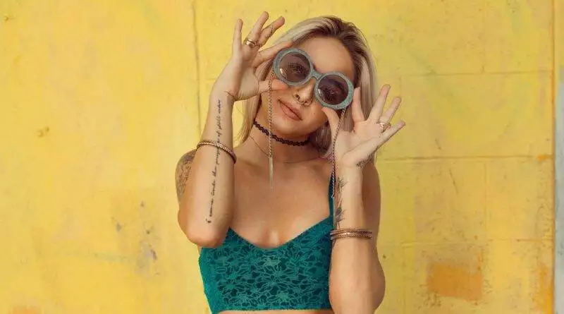 Eine Frau in einem trendigen grünen Top und Sonnenbrille posiert vor einer leuchtend gelben Wand.